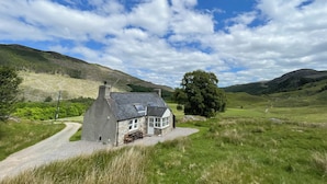 Une maison de campagne avec une façade en pierre, une allée en gravier et un grand arbre à proximité.