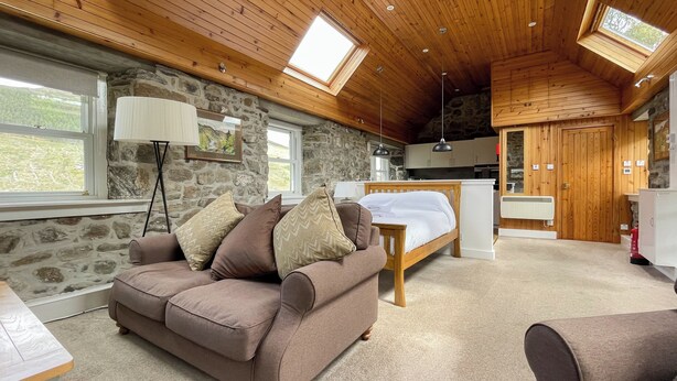 Une pièce chaleureuse avec un mur en pierre, un plafond en bois, un lit, un canapé et un petit coin cuisine.