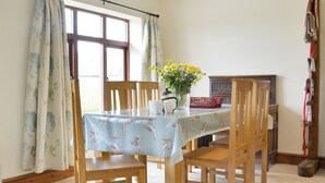 A dining area with a table covered by a tablecloth, wooden chairs, a vase with flowers, and a window with curtains. - The Granary - W43949 (East Tytherton, Chippenham)