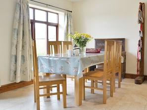 Ein Essbereich mit einem Tisch, der mit einer Tischdecke bedeckt ist, Holzstühlen, einer Vase mit Blumen und einem Fenster mit Vorhängen.