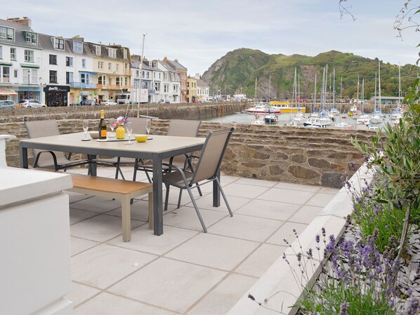 A patio with a table set for dining, overlooking a harbor with boats and a hillside in the background.
