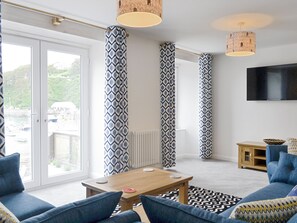 A modern living room with a large window, a wooden coffee table, blue sofas, patterned curtains, and a television.