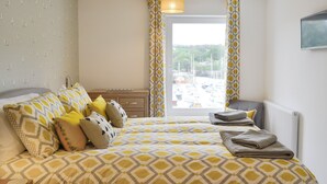 A neatly made bed with patterned bedding, a bedside table with a lamp, and a view of boats through a window. - 3 bedroom accommodation in Ilfracombe (Ilfracombe)
