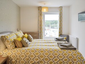 Een net opgemaakt bed met beddengoed met een patroon, een nachtkastje met een lamp, en een uitzicht op boten door een raam.