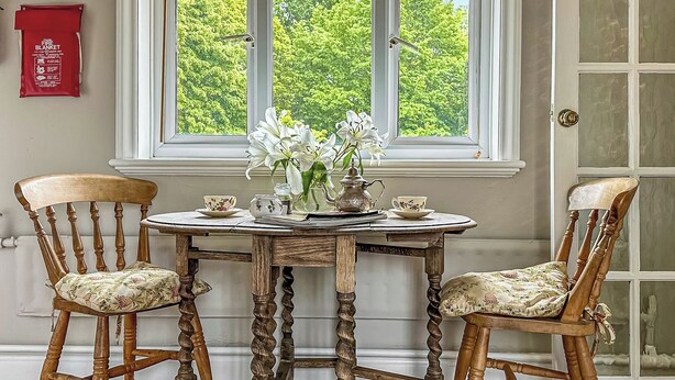 A room with a round wooden table, two chairs with floral upholstery, a vase with white flowers, and a window with a view of green trees. - 1 bedroom accommodation in Hollesley, near Woodbridge (Hollesley, near Woodbridge)