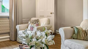A living room with a beige sofa, a white armchair, and a vase of white lilies. A door with a golden handle is visible in the background. - 1 bedroom accommodation in Hollesley, near Woodbridge (Hollesley, near Woodbridge)