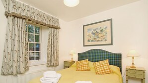Una cama bien hecha con ropa de cama amarilla y un cabecero de cuadros, dos lámparas de noche, un cuadro en la pared y una ventana con cortinas florales.