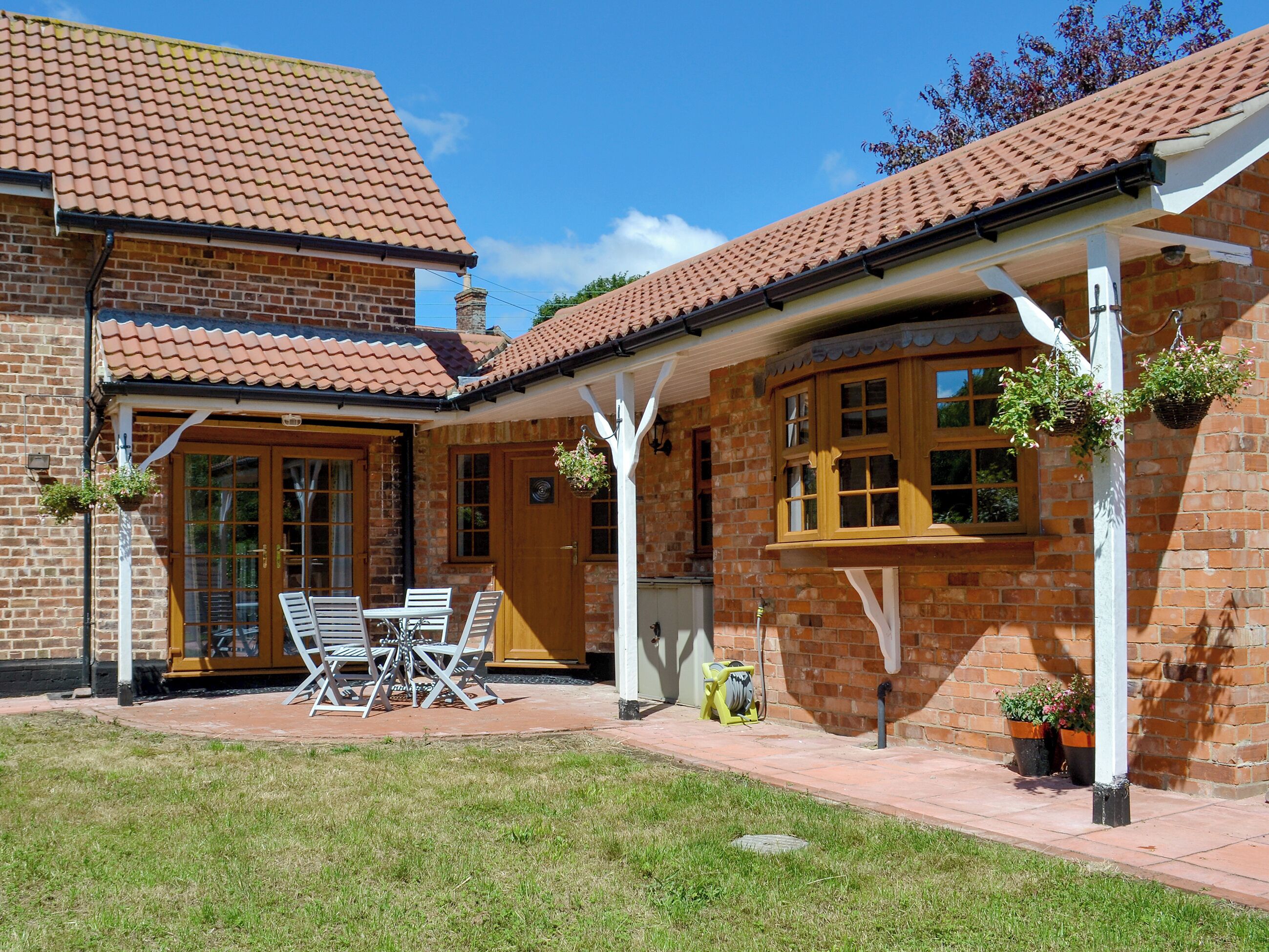 A brick house with a tiled roof, a covered patio with a table and chairs, and hanging flower baskets.