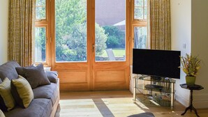 A living room with a sofa, a flat-screen TV on a stand, and a view through a large wooden door to a garden. - 2 bedroom accommodation in Sheringham (Sheringham)