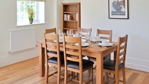 A dining room with a wooden table set for six, surrounded by six wooden chairs with black cushions. A bookshelf is against the wall, and a framed picture hangs above the table. - 2 bedroom accommodation in Sheringham (Sheringham)