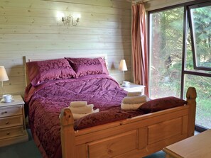 A wooden-paneled room with a bed, two bedside tables, a lamp, and a window with curtains. - Hartland Forest Golf & Leisure (Woolsery, near Bideford)