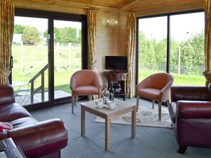 A living room with leather sofas, a coffee table, and a television. There are curtains, a window, and a view of a garden. - Hartland Forest Golf & Leisure (Woolsery, near Bideford)