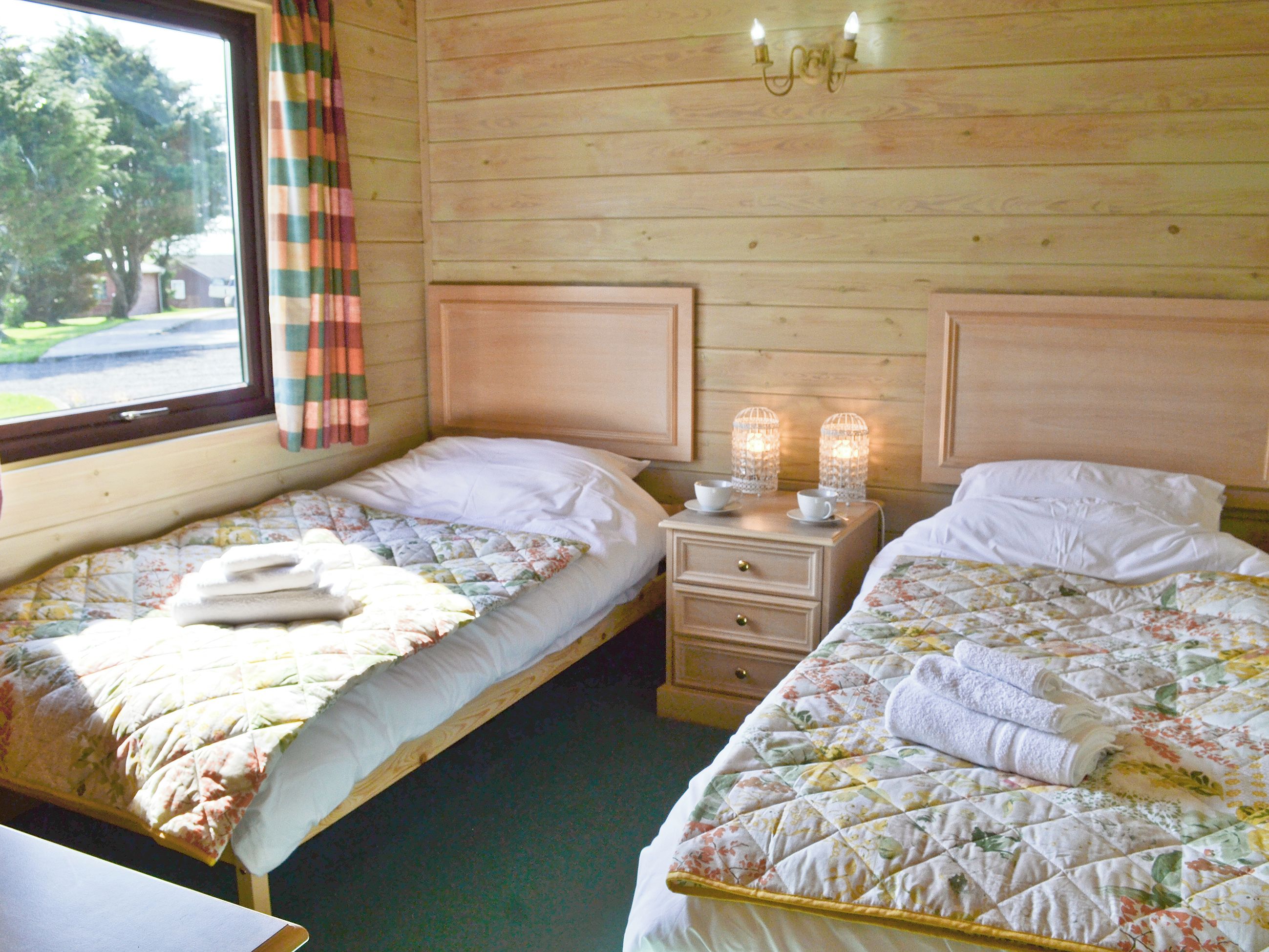 A room with two beds, each with a quilted bedspread and pillows, a nightstand with two lit candles, and a window with curtains.
