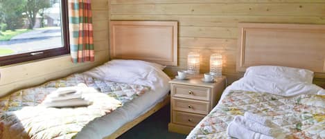Une chambre avec deux lits, chacun recouvert d’une courtepointe et doté d’oreillers, une table de chevet avec deux bougies allumées et une fenêtre avec des rideaux.
