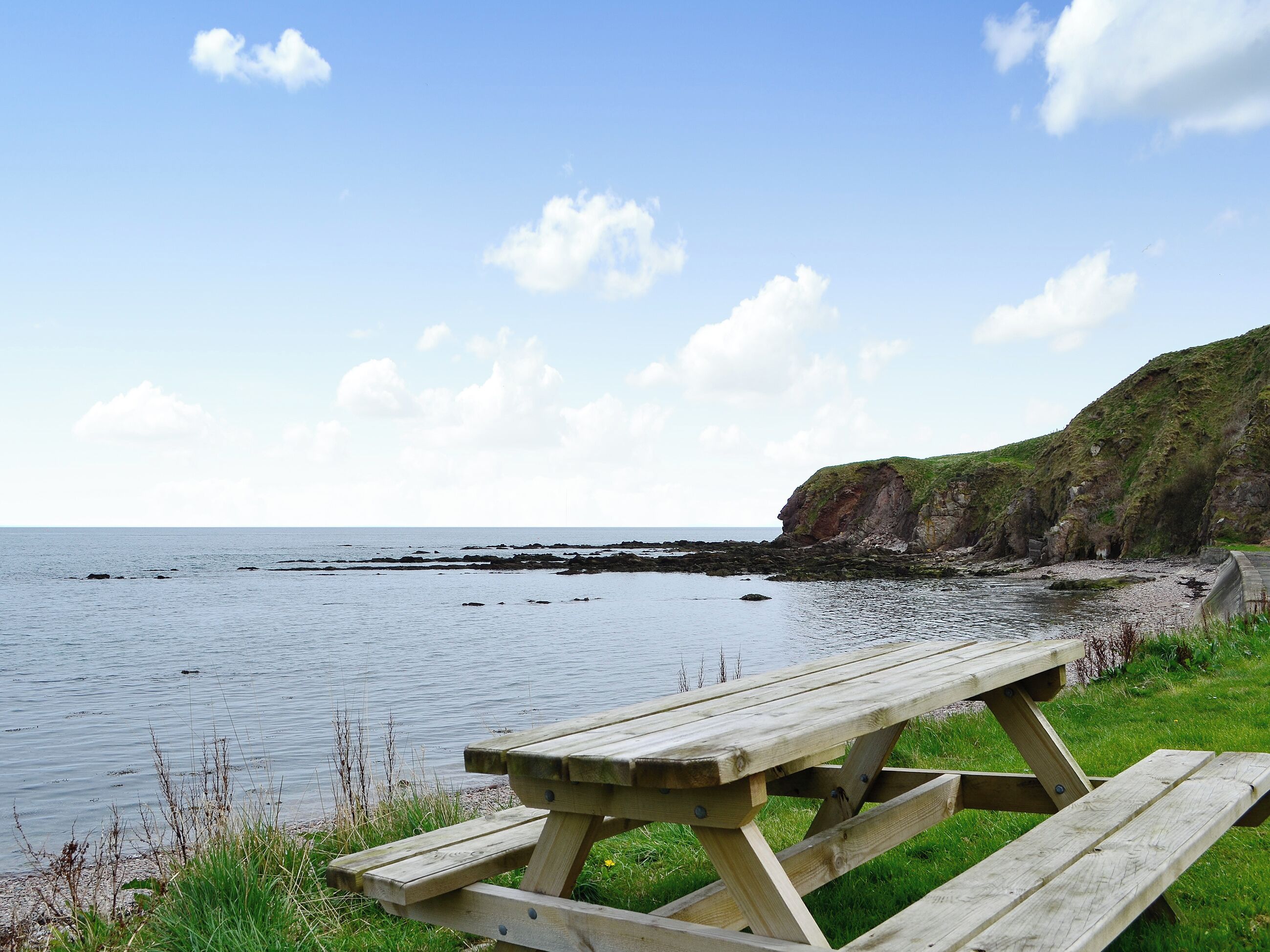 A wooden picnic table by a calm sea with a grassy area and a rocky cliff in the background.