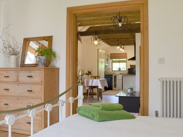A bedroom with a wooden dresser, a bed with green towels, a mirror, and a view into the kitchen.