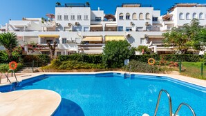 Une piscine à l’eau d’un bleu limpide, entourée d’une terrasse aux couleurs claires. Au fond, on aperçoit des bâtiments blancs dotés de balcons et quelques espaces verts.