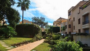Un sendero atraviesa un jardín cuidado con palmeras y arbustos, bordeado por edificios con balcones y tejado de tejas.
