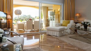 Amplia sala de estar con sofá, sillones, una mesa de centro de vidrio y televisor. Incluye un balcón con mobiliario exterior y vista a zonas verdes.