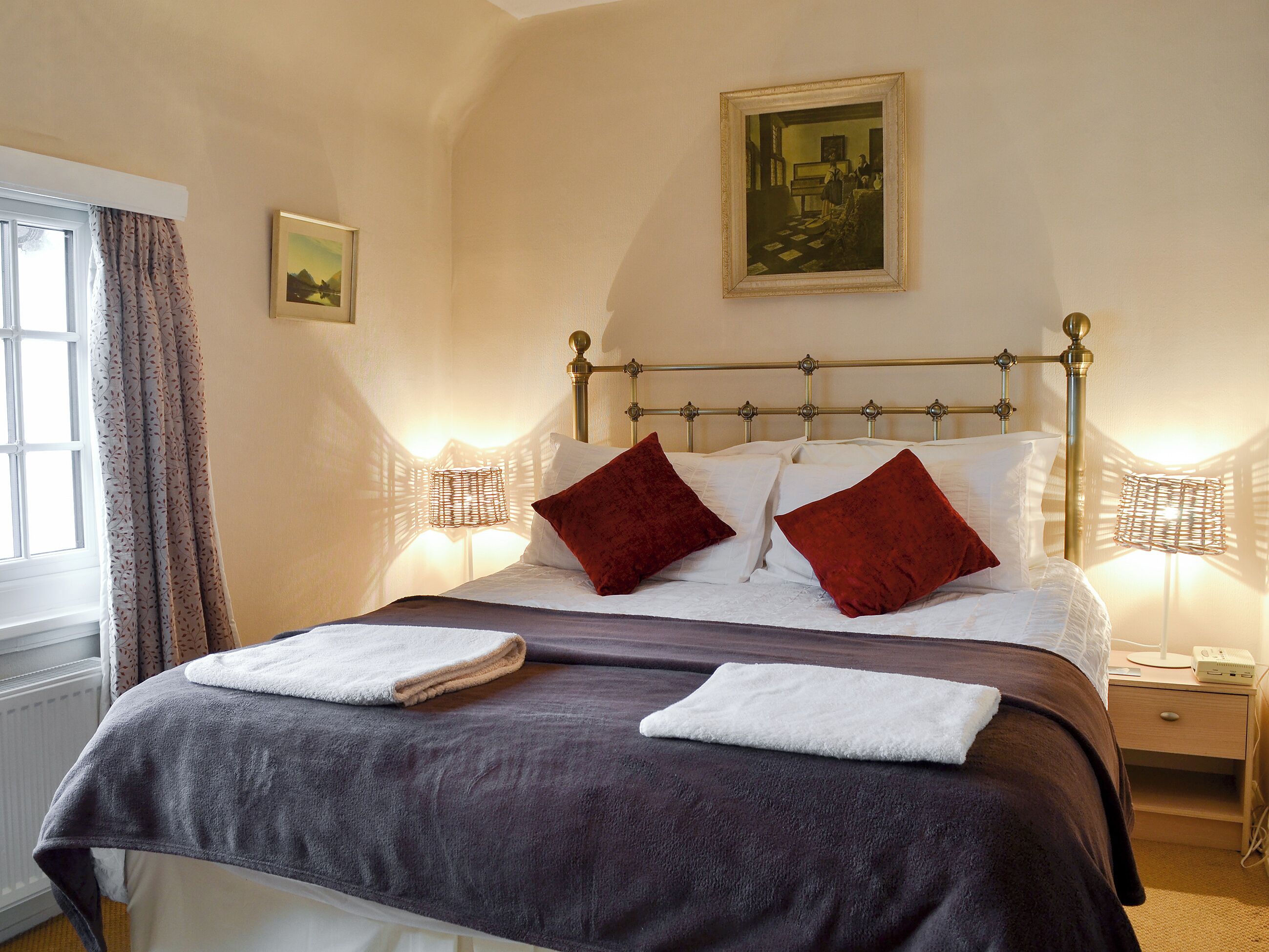 A neatly made bed with two white towels, a dark brown bedspread, and two red pillows. There is a framed picture on the wall above the bed, a window with curtains, and a bedside table with a lamp.