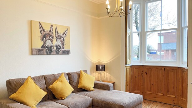 A living room with a sectional sofa, a lamp, and a painting of donkeys.