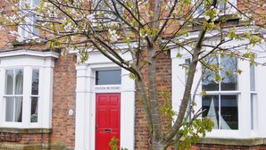A red door with a white window above it, a brick wall, and a small tree in front.