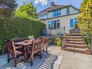 Un patio con mesa y sillas de madera, un pequeño jardín y una casa de dos plantas con una escalera visible.