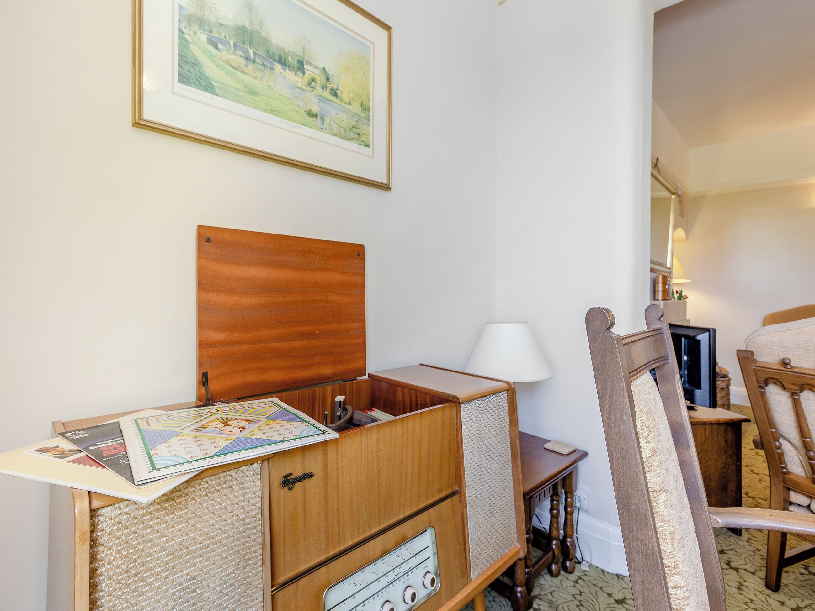 Ein Zimmer mit einem Holzkasten, einem kleinen Tisch mit Lampe, einem Sessel und einem gerahmten Bild an der Wand.