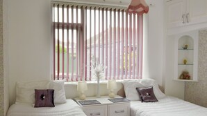 A bedroom with two beds, a window with pink blinds, bedside tables with lamps, and a small shelf with decorative items.