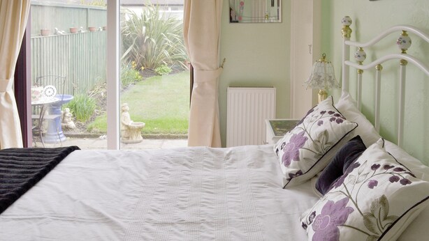 A bedroom with a bed, a window with curtains, a radiator, and a view of the garden.