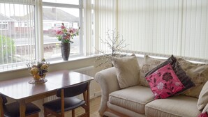 A living room with a beige sofa, a wooden dining table with chairs, a red rug, and a window with a view of houses and a street.