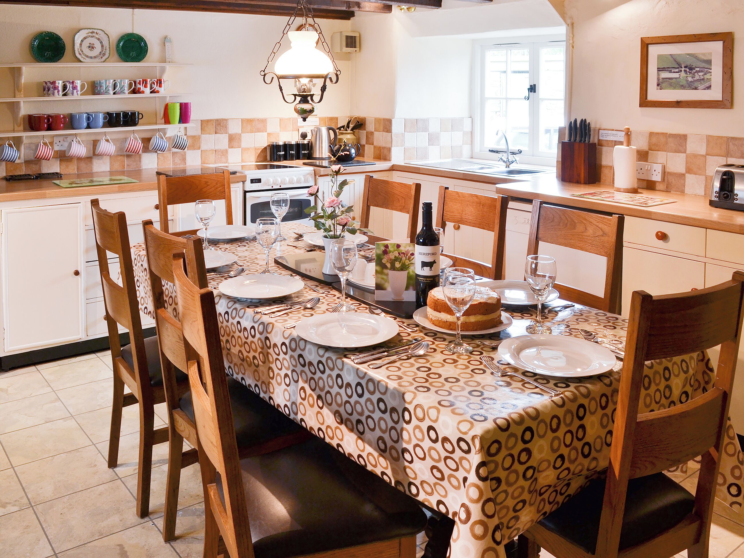 Een eetgedeelte met een tafel voor vier personen, stoelen, een kroonluchter en een keuken op de achtergrond.