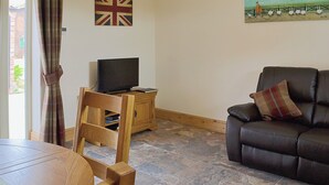 Ein Wohnzimmer mit Fernseher auf einer Holzkommode, einem braunen Ledersofa, einem karierten Kissen und einem Gemälde, das eine Bauernszene zeigt.
