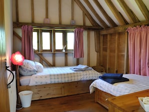 Una habitación acogedora con dos camas, vigas de madera, una ventana con cortinas, una lámpara de noche y un balde pequeño.