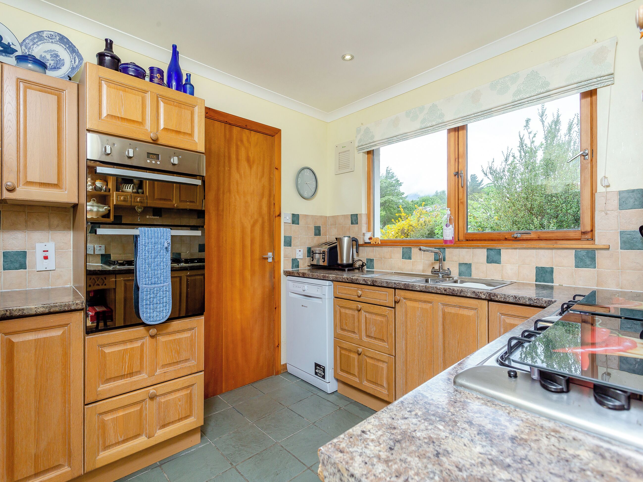 A kitchen with wooden cabinets, a built-in oven, a dishwasher, and a window with a view of greenery.