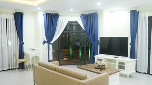 A modern living room with a beige sofa, a coffee table, a TV, and a glass door leading to a balcony. - QB Casa Residence (Dong Hoi)