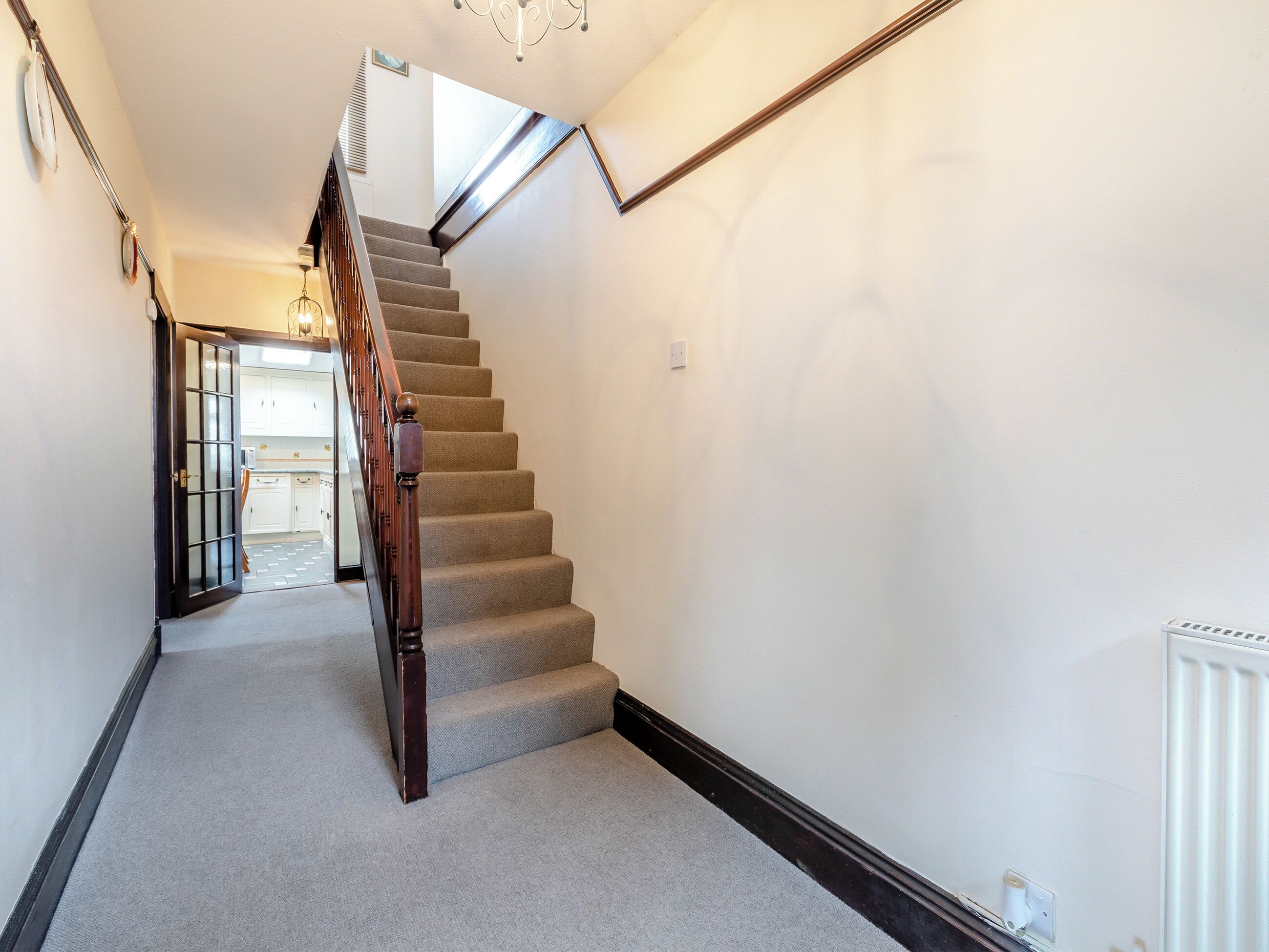 A carpeted staircase with wooden handrails, leading to a kitchen area with white cabinets and a visible light fixture.