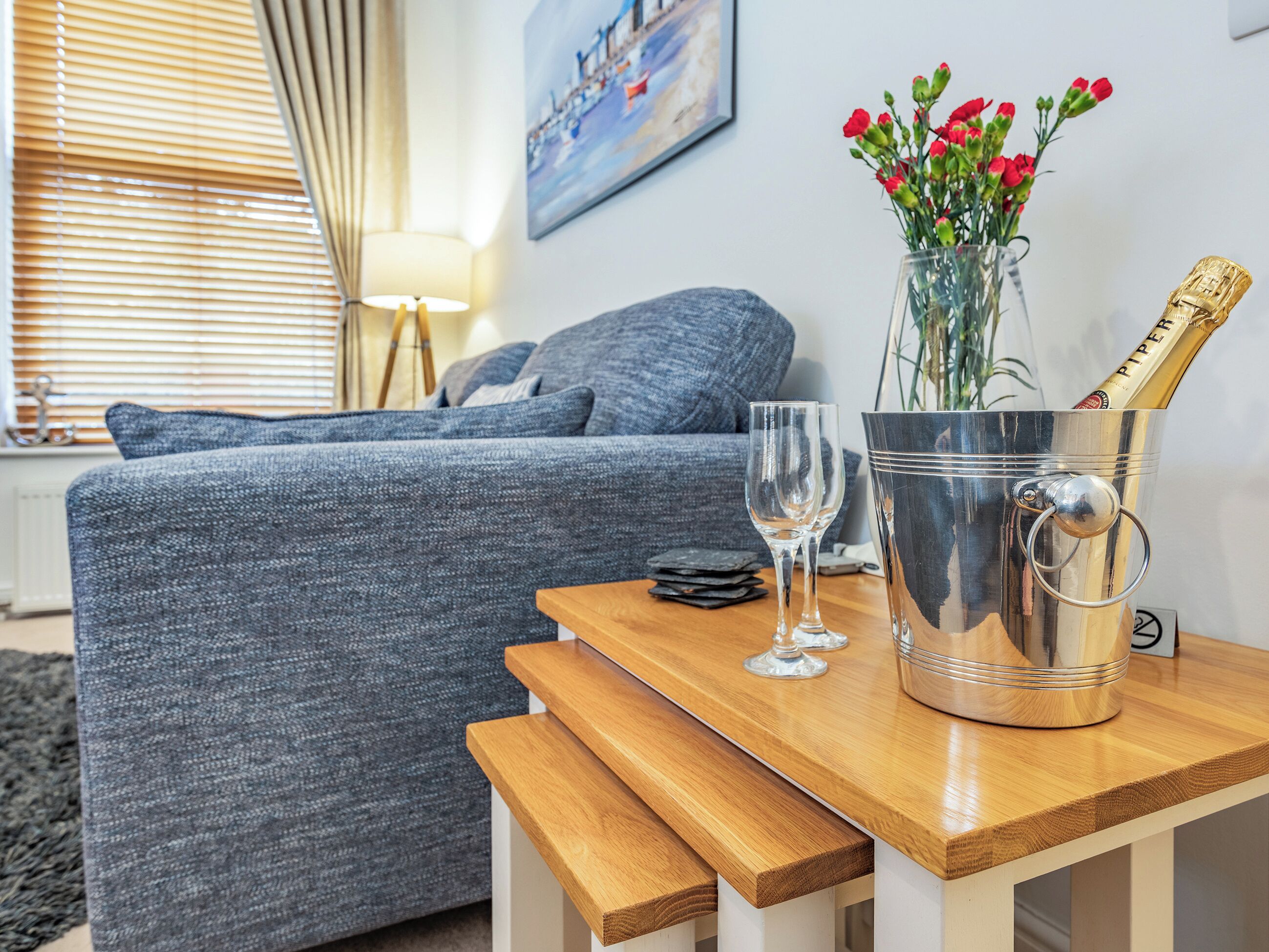 A modern living room with a grey sofa, a wooden coffee table, a metal champagne bucket, and a vase with red flowers.
