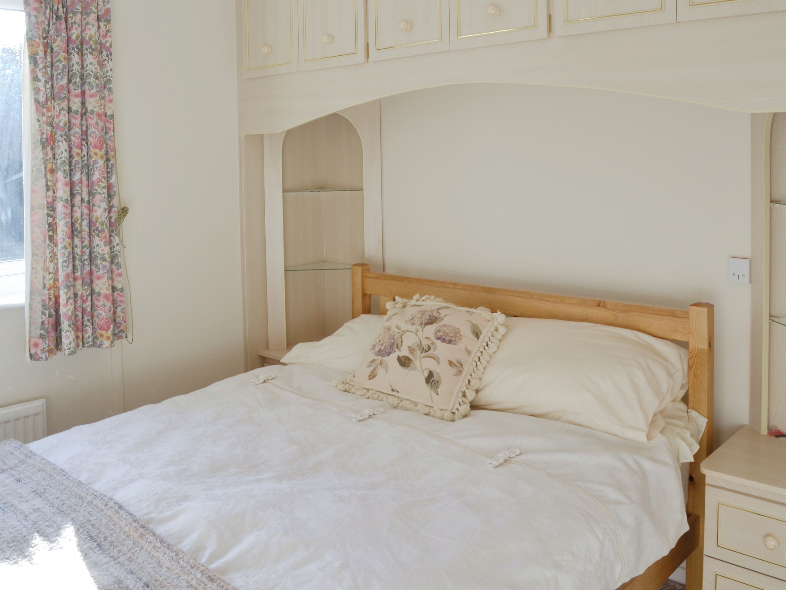 A bed with a wooden headboard, a floral pillow, and a white bedspread. There is a nightstand with a drawer and a small table beside the bed. A glass shelf is mounted on the wall above the bed.