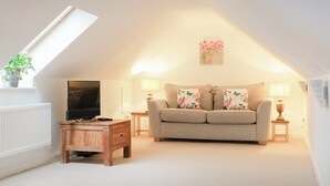 Ein gemütliches Wohnzimmer mit hohen Decken, einer Couch mit floralen Kissen, einem Holztisch, einem Fernseher und einer Pflanze auf einem Fensterbrett.