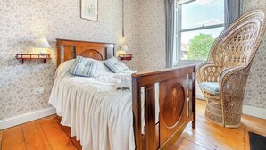 Un dormitorio con cama de madera, una silla de mimbre, una mesita de noche con lámparas y papel tapiz floral.