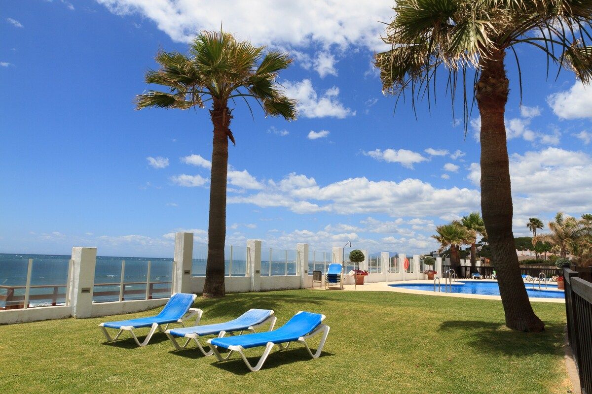 Zona junto a la piscina con sillas de descanso y palmeras, con vistas a un cielo azul despejado y una masa de agua.