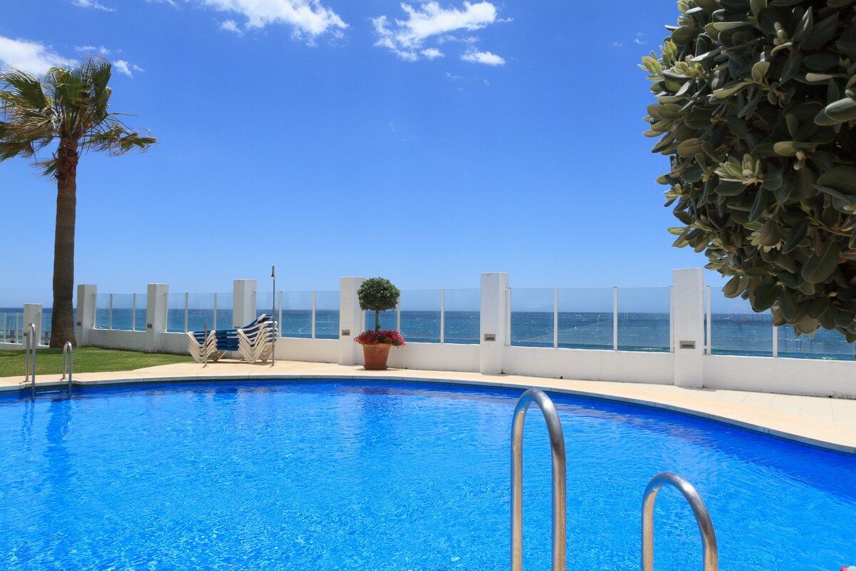 A swimming pool with a view of the sea, surrounded by a white fence, and a palm tree.