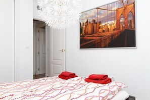 Une chambre d’hôtel avec un lit, une porte blanche, un tableau représentant un paysage urbain encadré et un lustre blanc.
