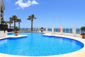 A swimming pool with a curved edge, surrounded by potted plants and lounge chairs, with a view of the sea and buildings in the background.