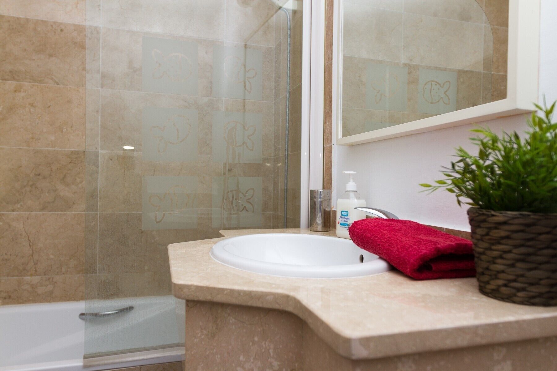 A bathroom with a beige marble sink, a red towel, a potted plant, and a glass shower enclosure.