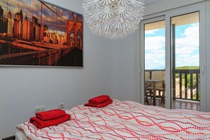 A hotel room with a bed, a cityscape painting, a chandelier, and a sliding glass door to a balcony.