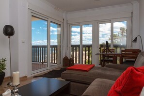 Un salon avec un canapé, une table basse et un coussin rouge. Il y a un balcon avec vue sur des palmiers et une terrasse en bois.
