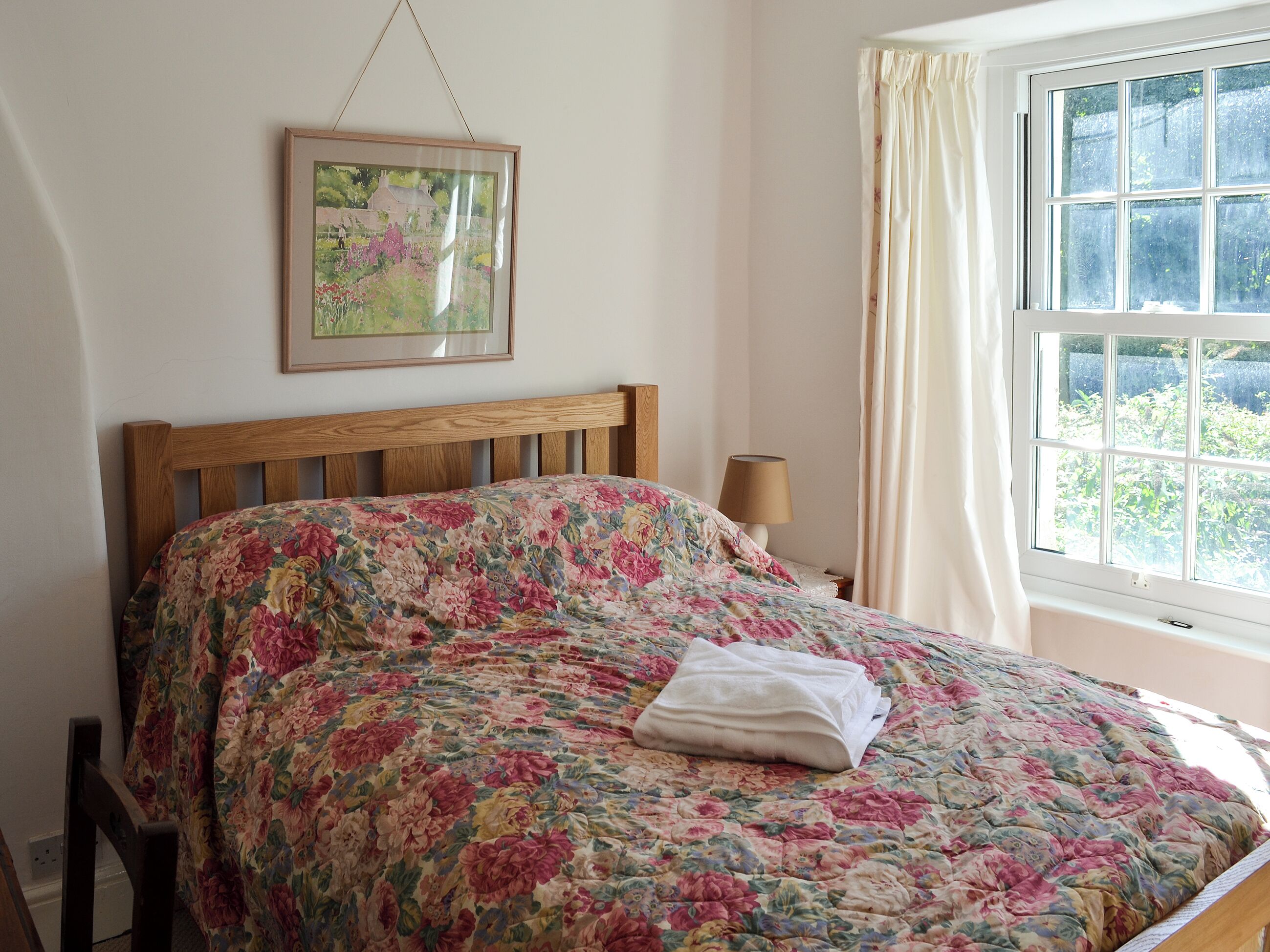 Een slaapkamer met een houten bed, bloemenbeddengoed, een bedlampje en een raam met gordijnen.