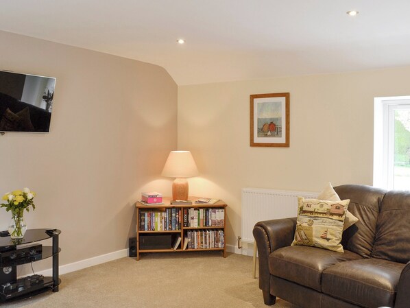 A living room with a brown leather sofa, a bookshelf with various books, a lamp, a framed picture on the wall, and a television mounted on the wall.